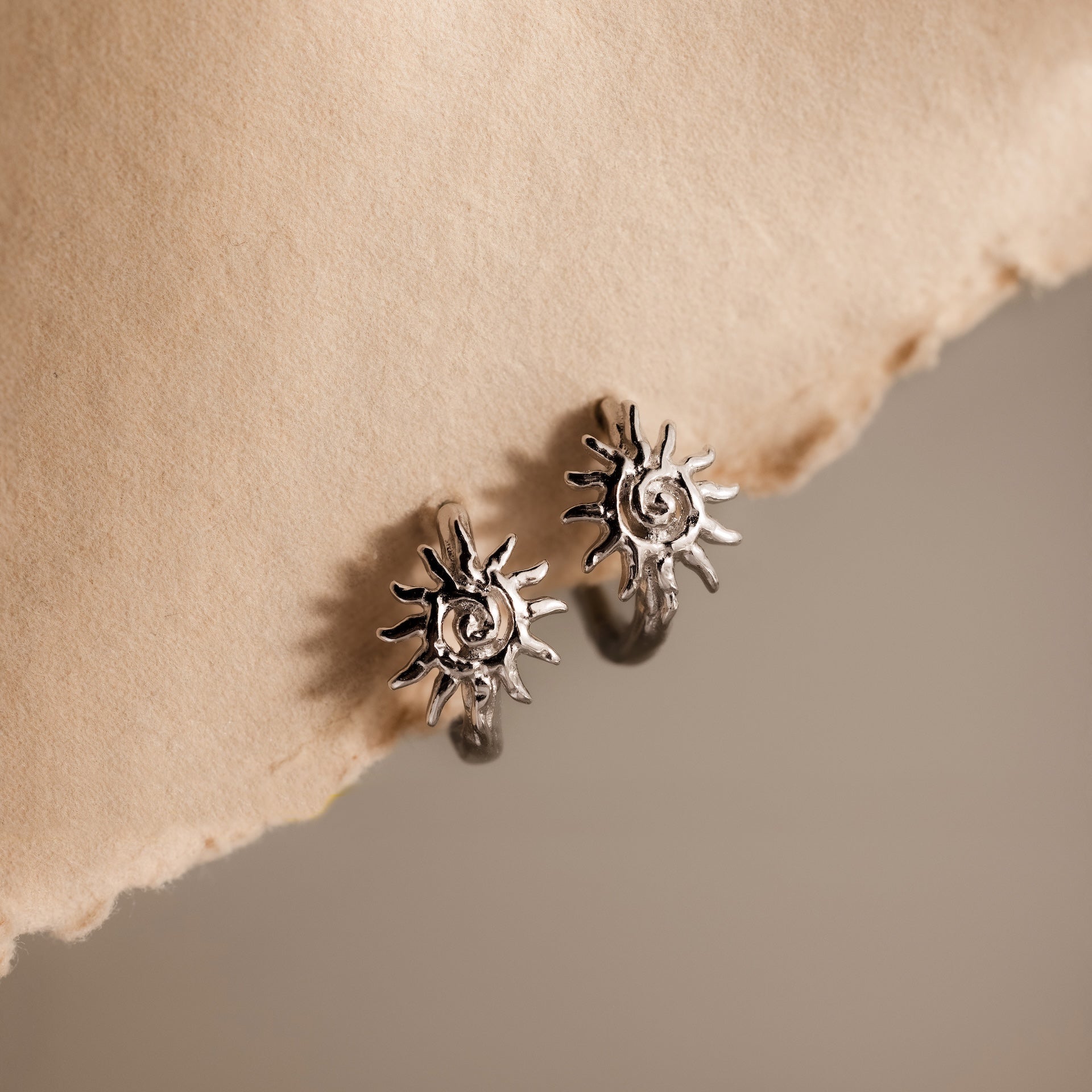 Celestial Sun Huggies in Silver, featuring a radiant sunburst design, are displayed on textured beige paper—perfect statement jewelry for any occasion.