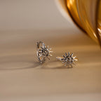 Celestial Sun Huggies in Silver, featuring sun-shaped designs, are displayed on a light surface near a golden accent—perfect for pairing with bold statement jewelry like sunburst earrings.