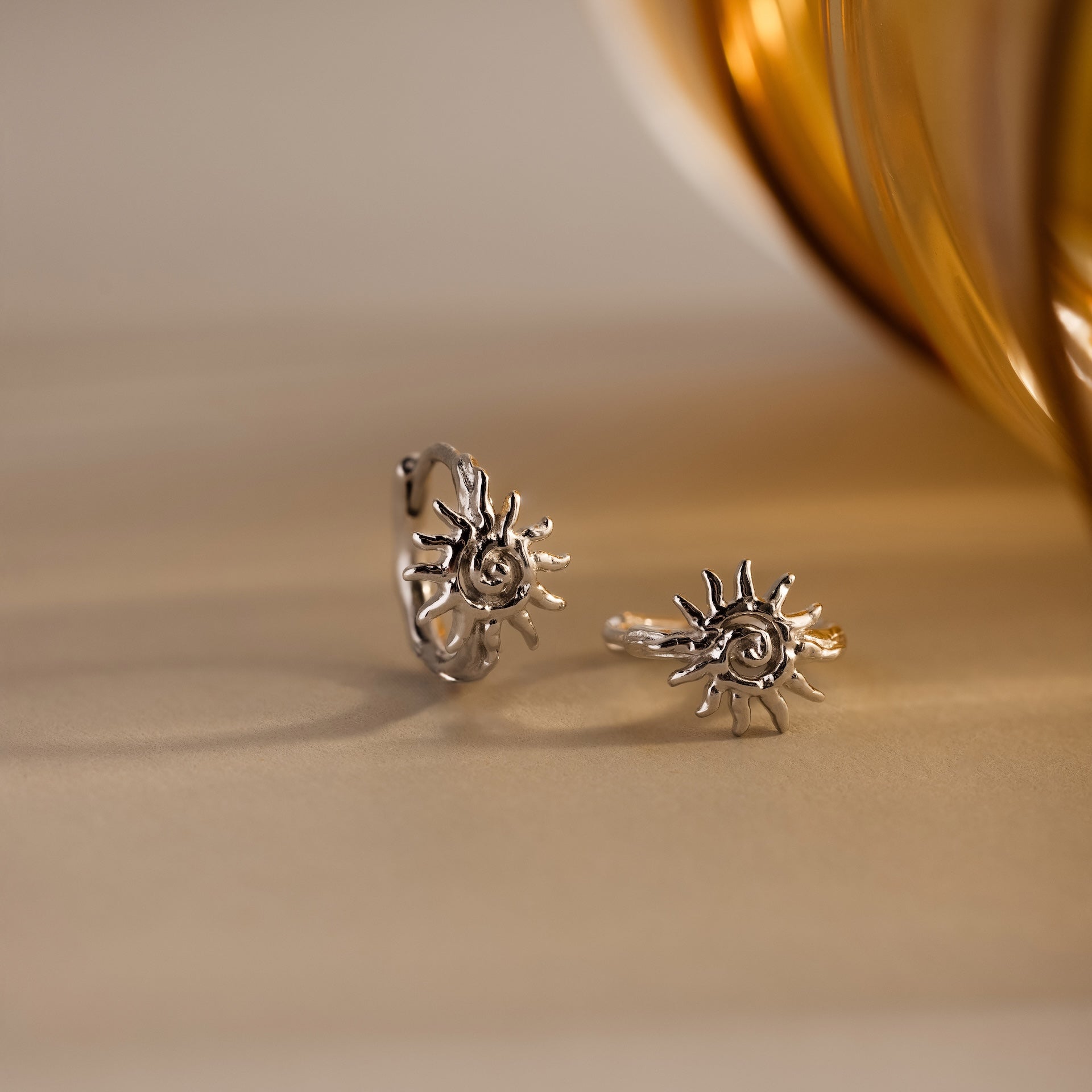 Celestial Sun Huggies in Silver, featuring sun-shaped designs, are displayed on a light surface near a golden accent—perfect for pairing with bold statement jewelry like sunburst earrings.