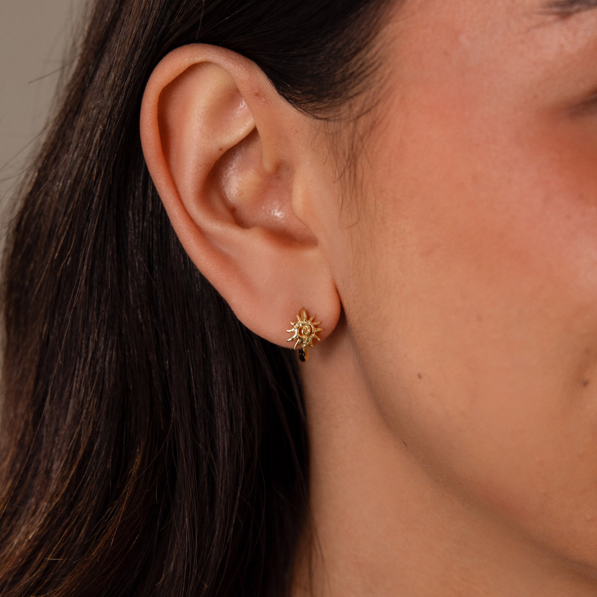 Close-up of a womans ear wearing Celestial Sun Huggies—gold sun-shaped earrings with a dangling detail, ideal for statement jewelry lovers.