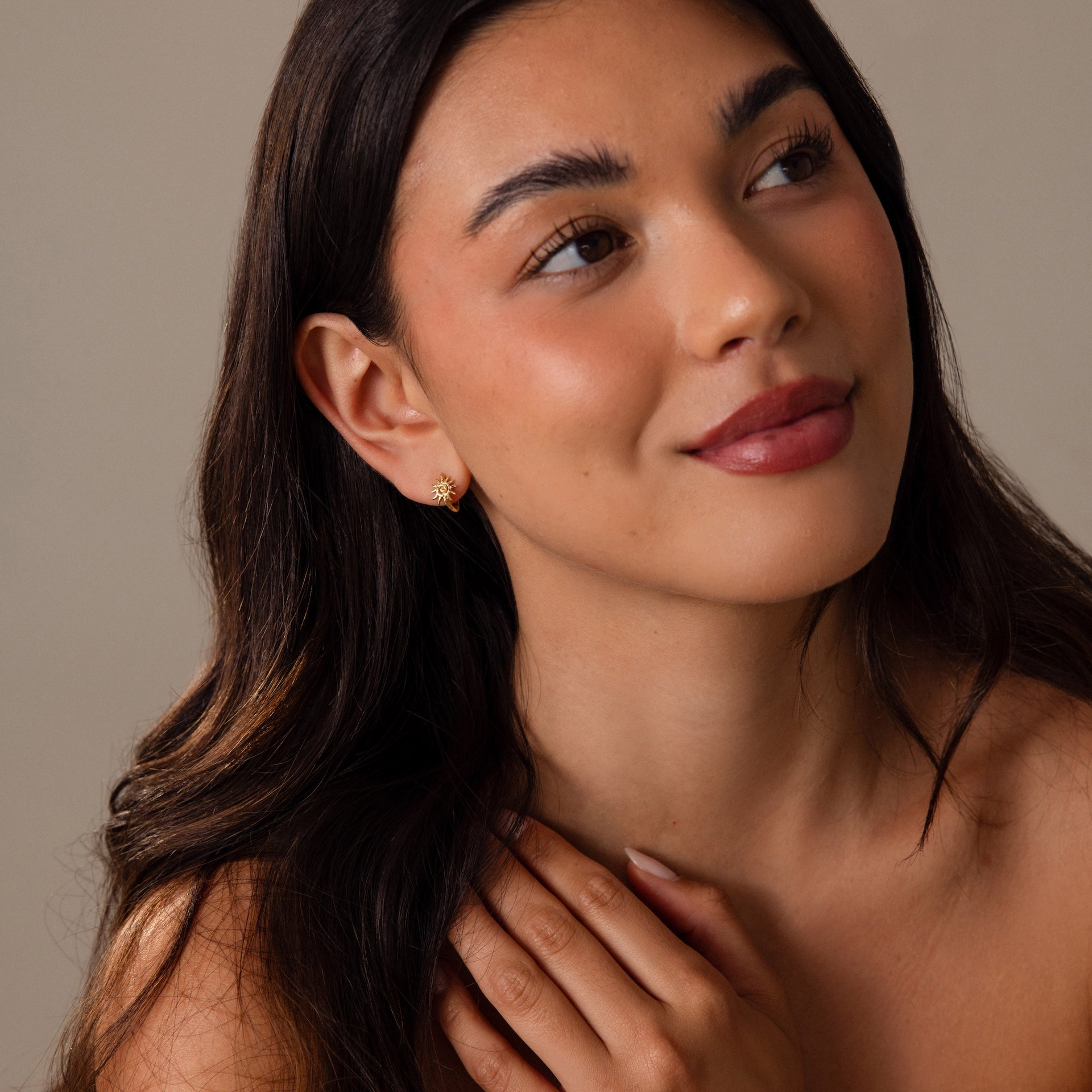 A woman with long dark hair smiles softly, wearing Celestial Sun Huggies in 18K Gold—these sunburst earrings add statement shine to her look.