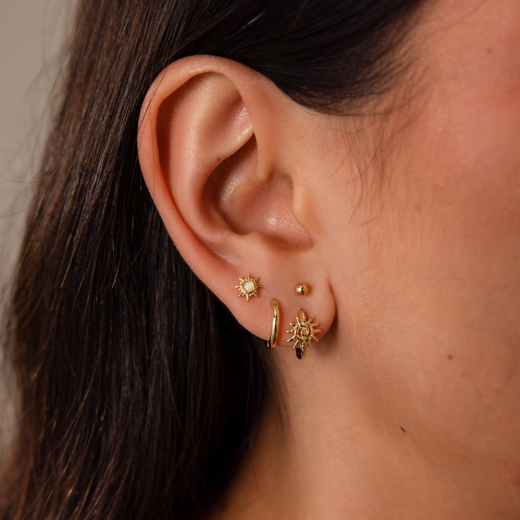 Close-up of an ear wearing statement jewelry: two sunburst earrings and Celestial Sun Huggies in 18K Gold with a sun pendant.