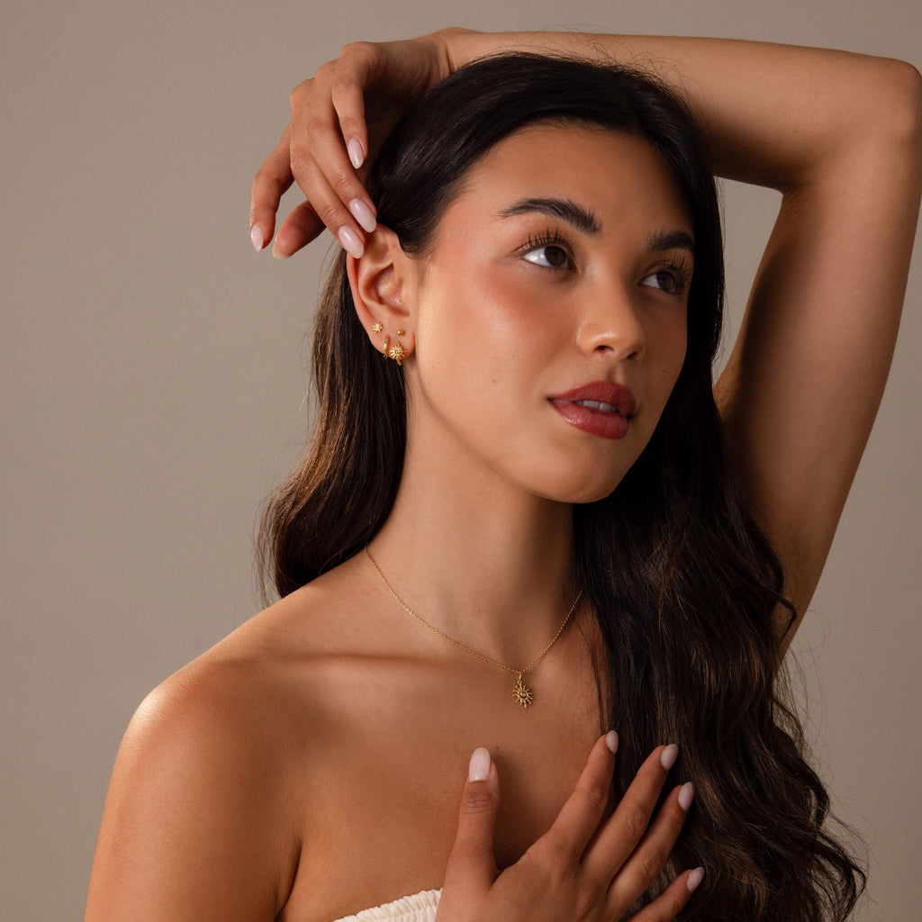 Woman with long dark hair poses with hand near head, wearing a strapless top and Celestial Sun Huggies in 18K Gold, set against a neutral background.