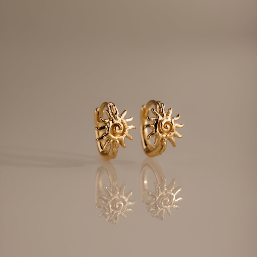 Celestial Sun Huggies in 18K Gold feature sun-shaped designs and are perfect statement earrings for any occasion, shown reflected on a smooth, neutral surface.