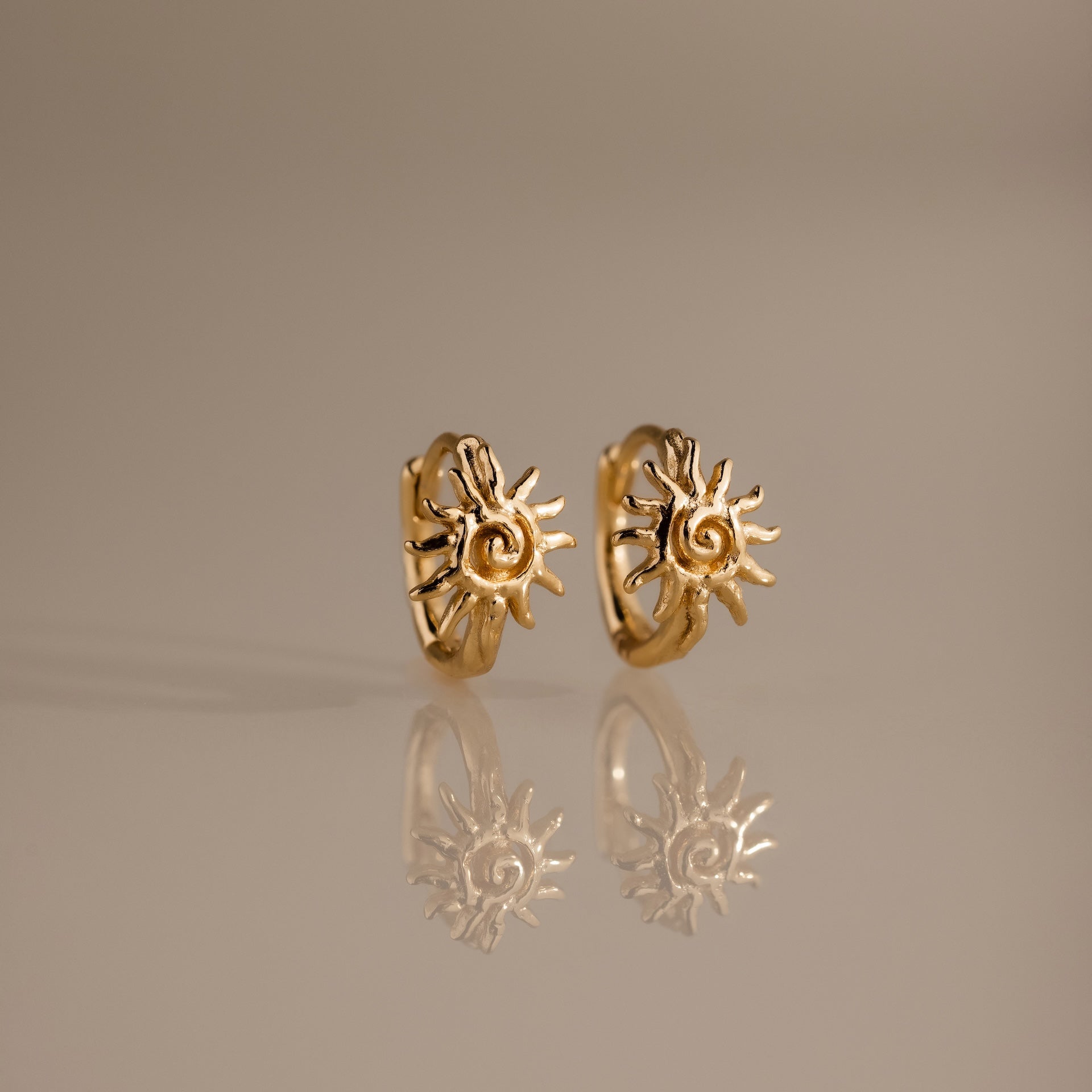 Celestial Sun Huggies in 18K Gold feature sun-shaped designs and are perfect statement earrings for any occasion, shown reflected on a smooth, neutral surface.