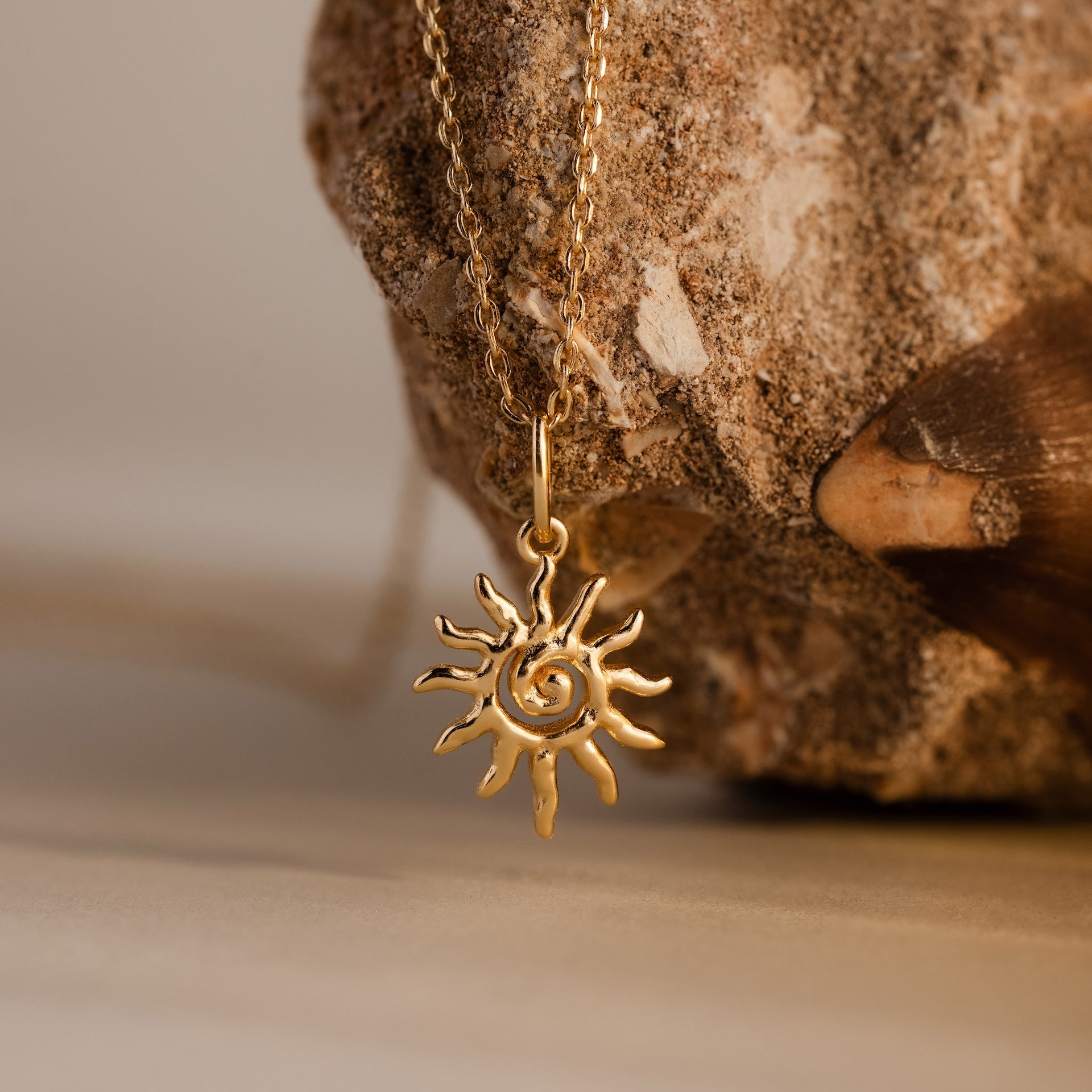 Celestial Sun Necklace in 18K Gold: A striking sunburst pendant statement necklace showcased on textured rock and shell.