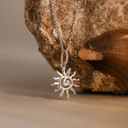 The Celestial Sun Necklace, featuring a bold sunburst pendant, is shown on a rock and beside a brown seashell—an ideal statement piece.