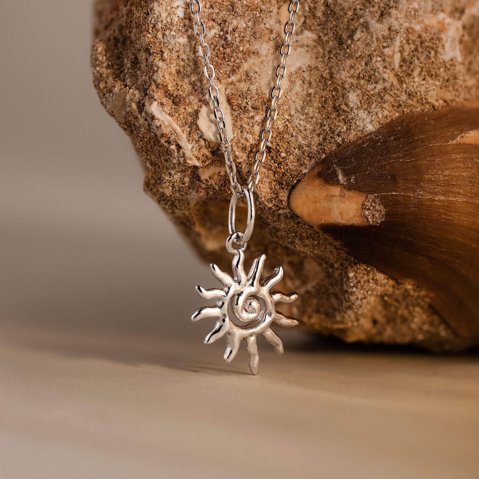 The Celestial Sun Necklace, featuring a bold sunburst pendant, is shown on a rock and beside a brown seashell—an ideal statement piece.