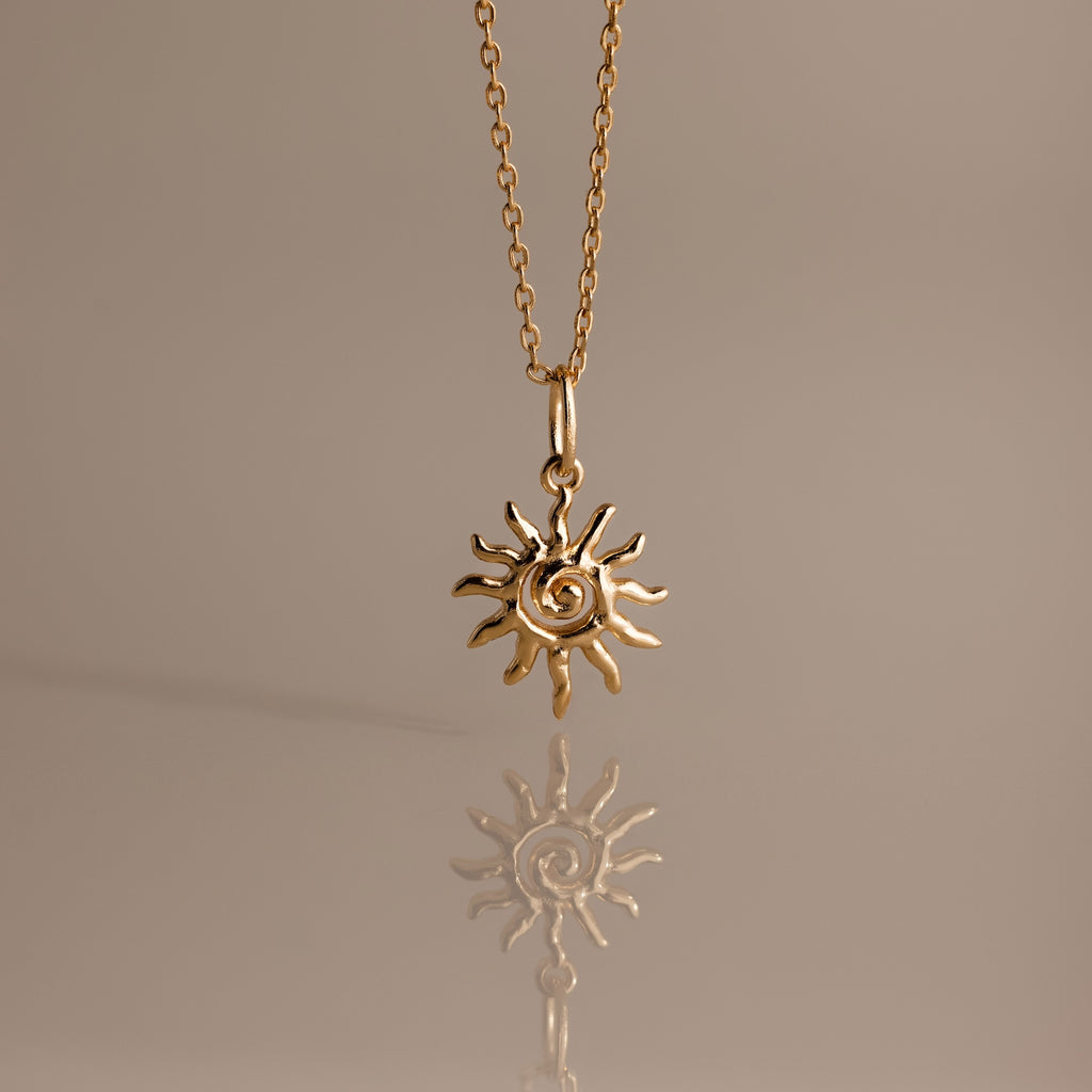 The Celestial Sun Necklace in 18K Gold features a swirling sunburst pendant, making a striking statement against any neutral background with its brilliant reflection below.