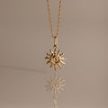 The Celestial Sun Necklace in 18K Gold features a swirling sunburst pendant, making a striking statement against any neutral background with its brilliant reflection below.