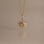 The Celestial Sun Necklace in 18K Gold features a swirling sunburst pendant, making a striking statement against any neutral background with its brilliant reflection below.