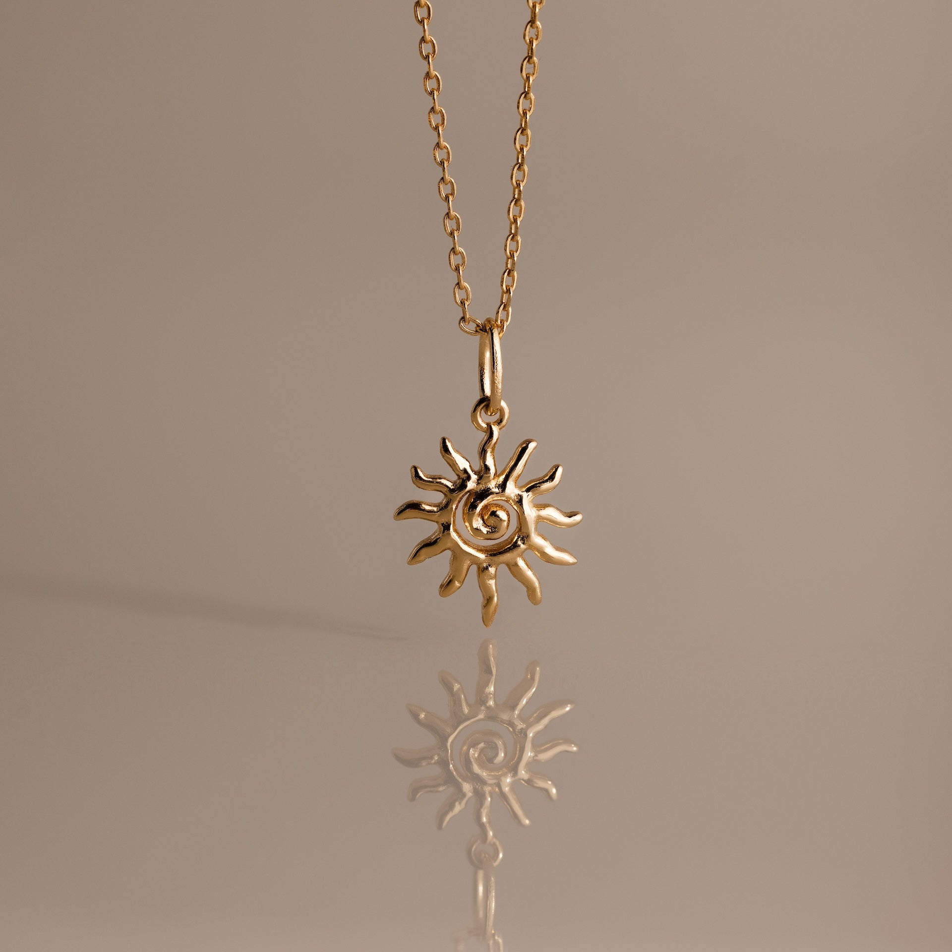The Celestial Sun Necklace in 18K Gold features a swirling sunburst pendant, making a striking statement against any neutral background with its brilliant reflection below.