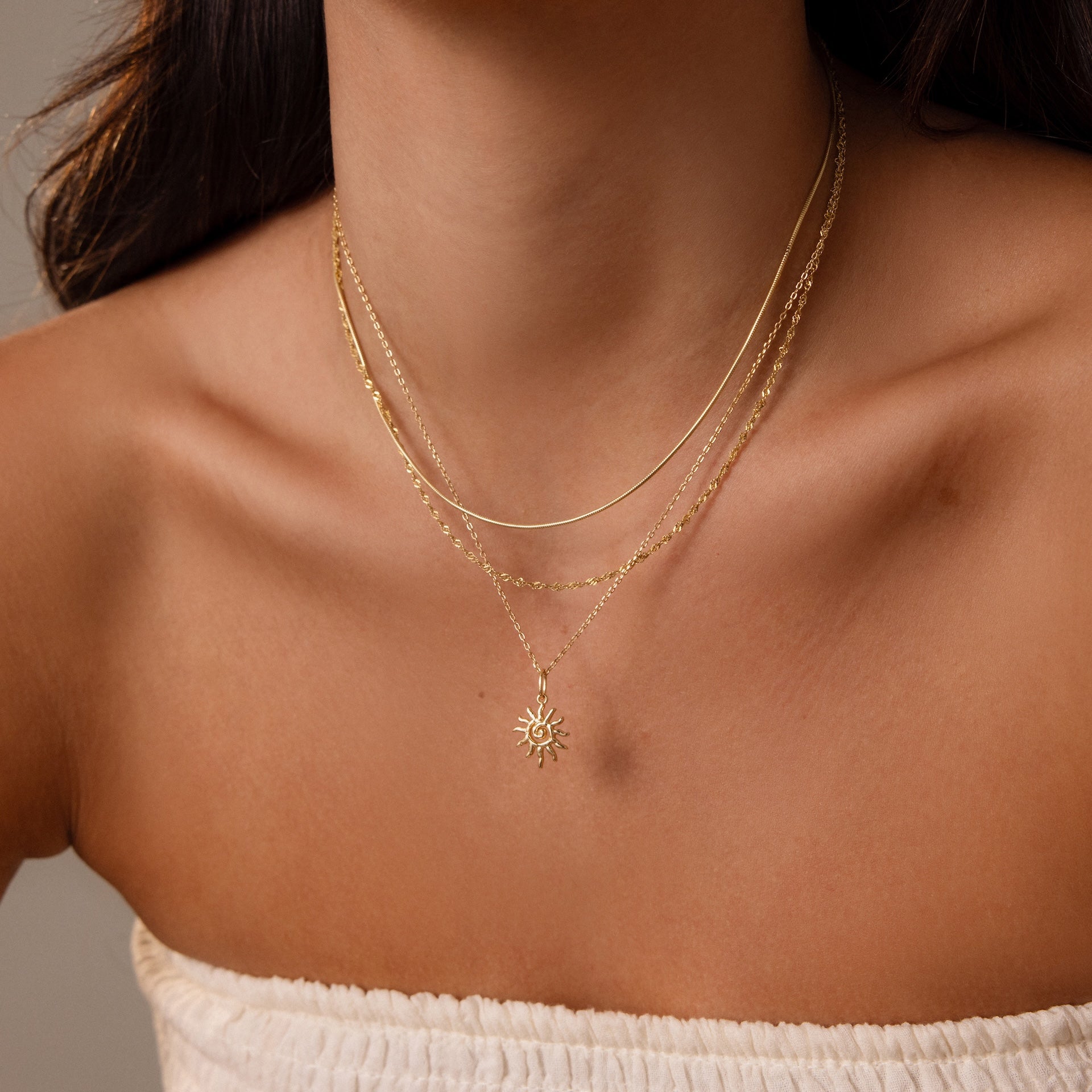 Woman wearing the Celestial Sun Necklace in 18K Gold with a sunburst pendant, layered with gold necklaces, and paired with a white off-shoulder top.