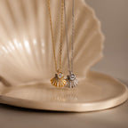 The Diamond Seashell Charm Necklace, featuring a gold and a silver shell pendant with gemstones, rests on a ceramic shell dish—coastal jewelry that radiates seaside elegance.