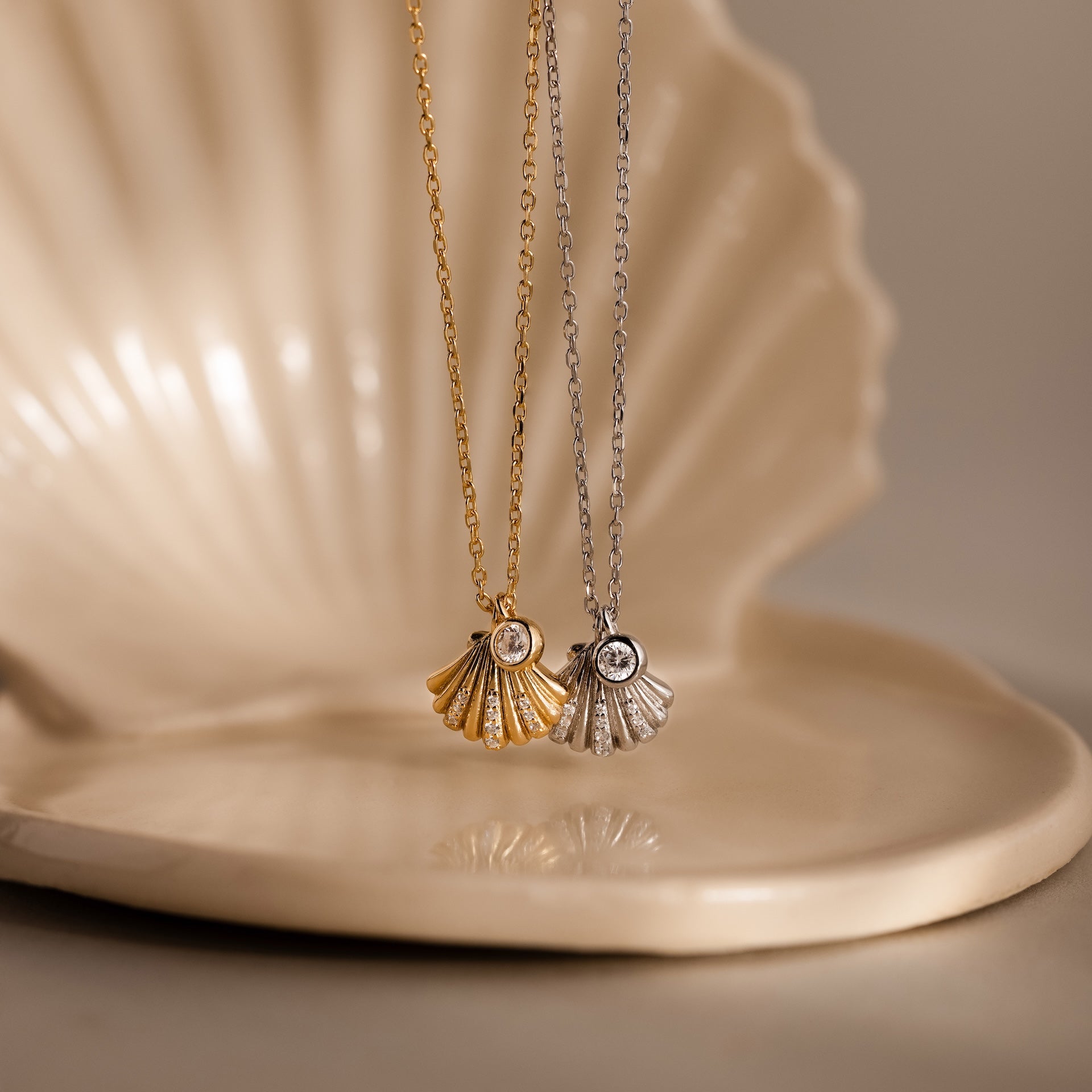 The Diamond Seashell Charm Necklace, featuring a gold and a silver shell pendant with gemstones, rests on a ceramic shell dish—coastal jewelry that radiates seaside elegance.