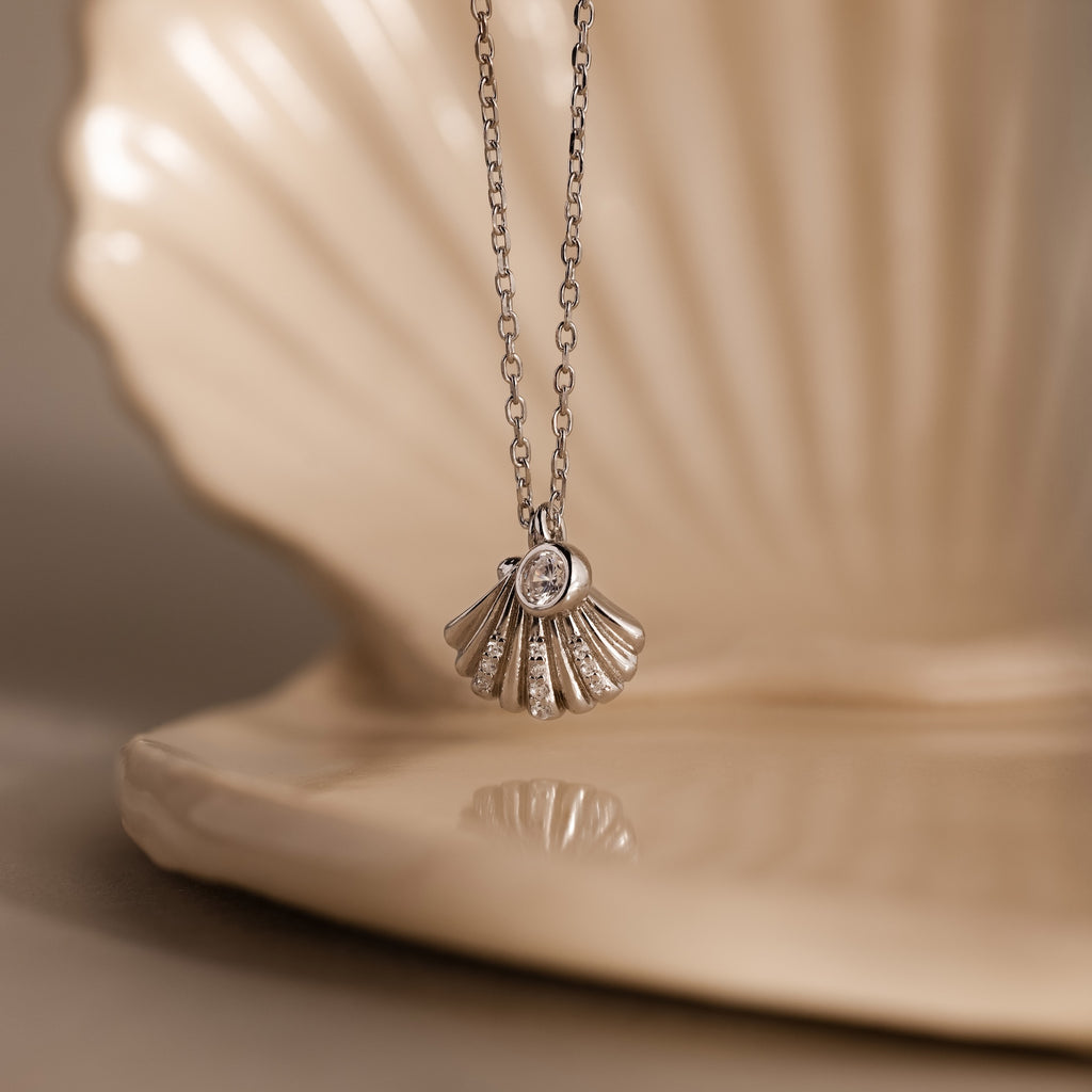 Diamond Seashell Charm Necklace in Silver, elegantly displayed before a large cream-colored shell.