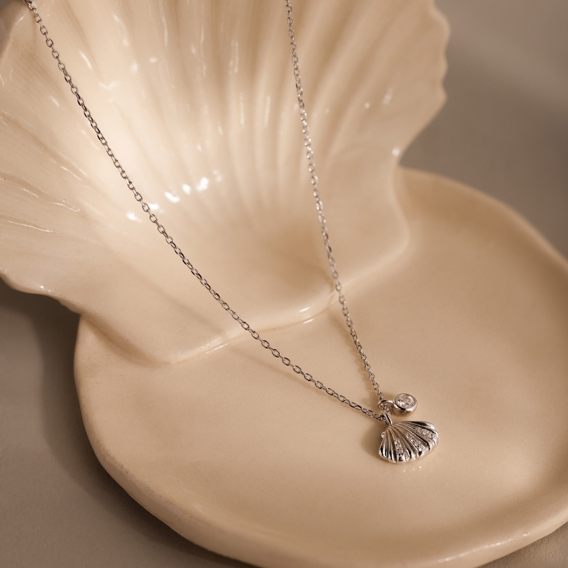 The Diamond Seashell Charm Necklace features a silver shell pendant with a diamond, elegantly displayed on a cream ceramic shell dish.