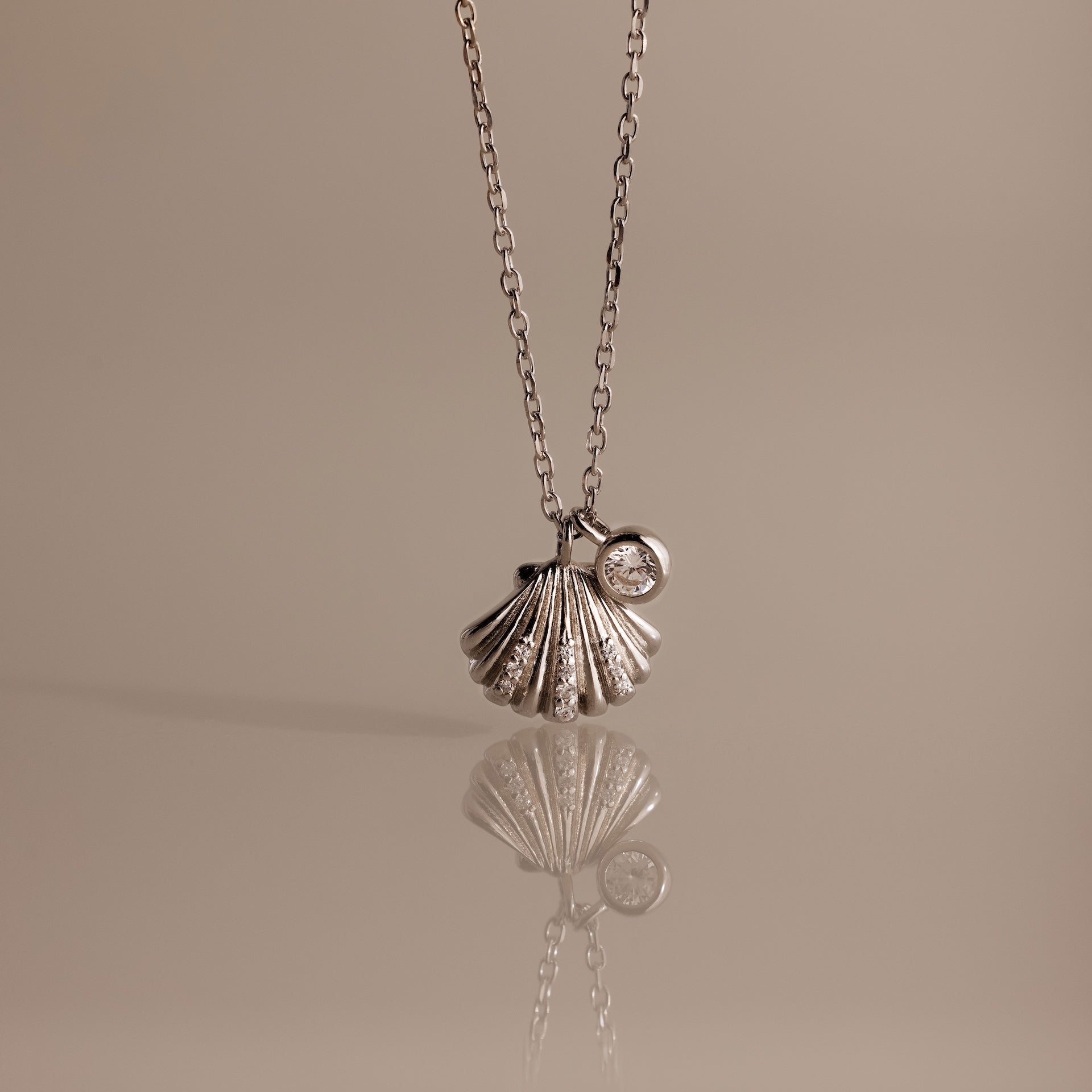 The Diamond Seashell Charm Necklace in Silver features a shell pendant and a small round gemstone, displayed on a neutral background—an elegant accessory to elevate any look.
