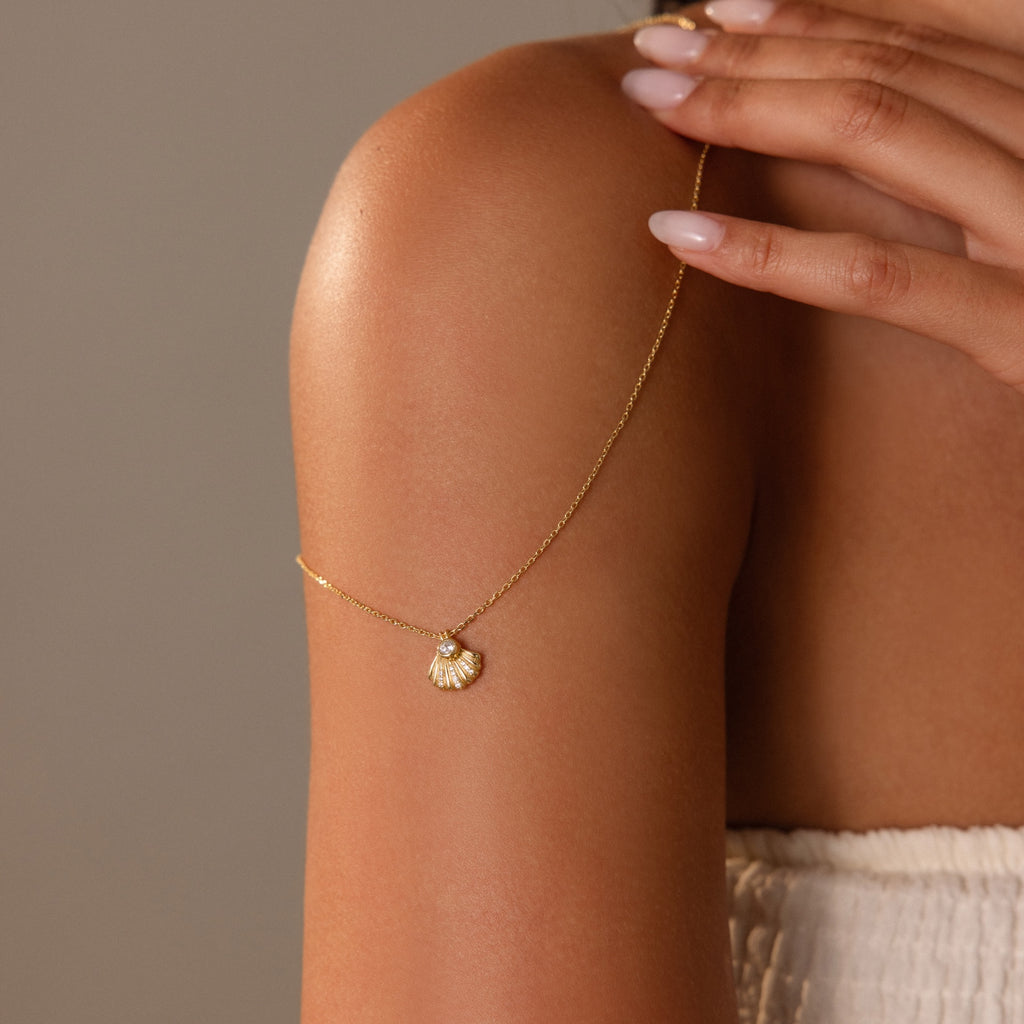 The Diamond Seashell Charm Necklace in 18K Gold drapes over a womans bare shoulder, her manicured nails highlighting the refined elegance of this stunning coastal-inspired piece.