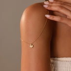 The Diamond Seashell Charm Necklace in 18K Gold drapes over a womans bare shoulder, her manicured nails highlighting the refined elegance of this stunning coastal-inspired piece.