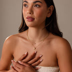 A woman in a strapless top gently touches her chest, showcasing layered gold necklaces, including the Diamond Seashell Charm Necklace in 18K Gold.