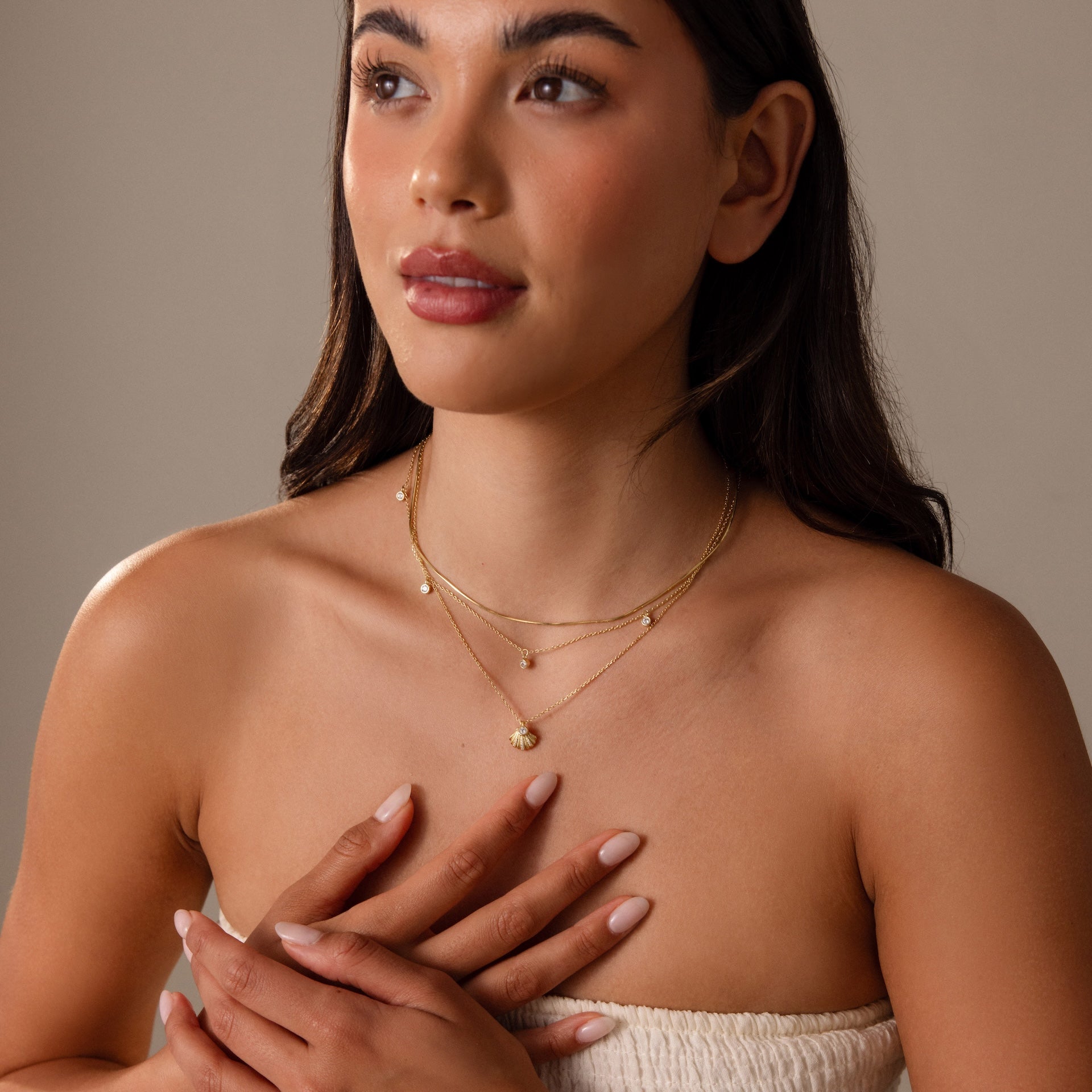 A woman with long dark hair wears a strapless top and layered gold necklaces, featuring the Diamond Seashell Charm Necklace, hands clasped near her chest.