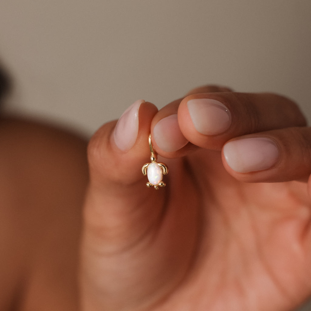 A hand with manicured nails holds the Opal Sea Turtle Charm, a small gold earring featuring a white oval stone and elegant ocean-inspired design, against a neutral background.