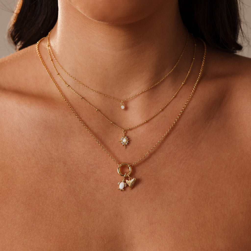 A woman wears three layered gold necklaces with small gemstone pendants and an Opal Sea Turtle Charm for an elegant, seaside-inspired look.