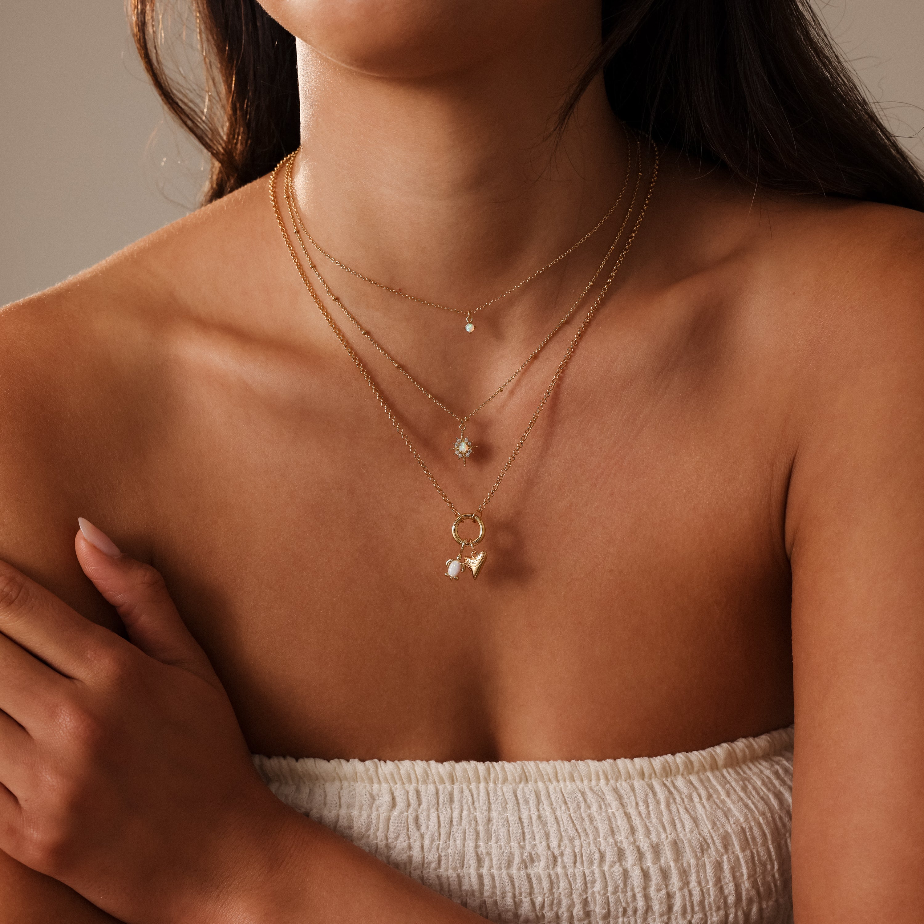 A woman wears the Opal Sea Turtle Charm layered with gold necklaces and a white textured strapless top for an ocean-inspired look.