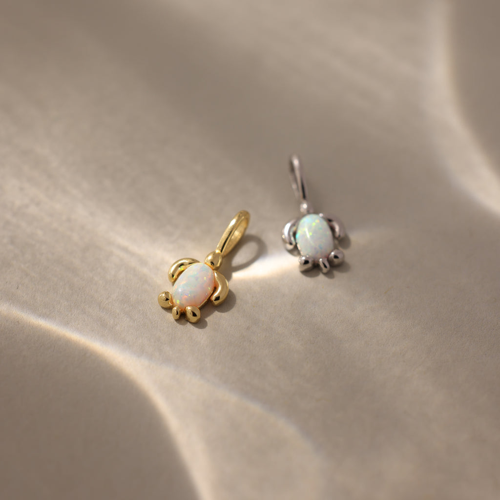 Two small Opal Sea Turtle Charms, one gold and one silver, are displayed on a beige surface—a perfect ocean-inspired accessory.