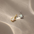 Two small Opal Sea Turtle Charms, one gold and one silver, are displayed on a beige surface—a perfect ocean-inspired accessory.