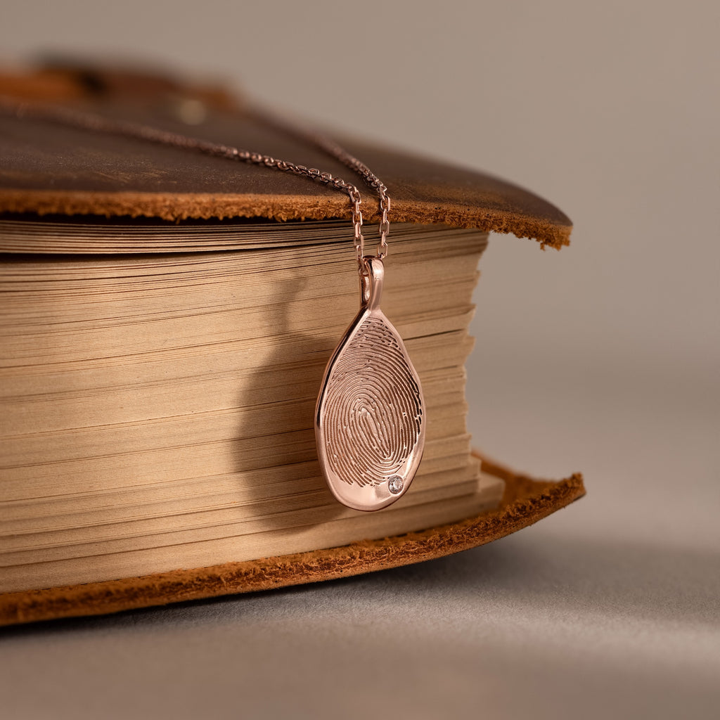 The Organic Fingerprint Teardrop Necklace in Rose Gold rests on an old, closed book—a timeless keepsake that captures cherished memories with its unique birthstone touch.