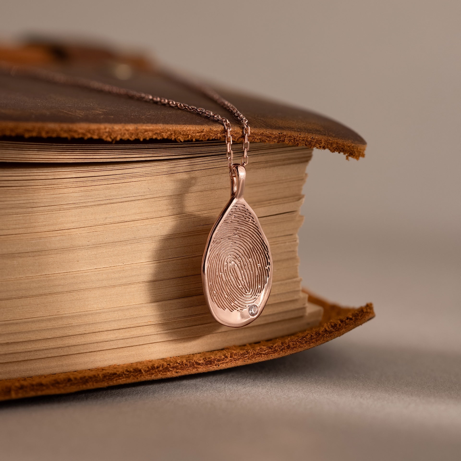 The Organic Fingerprint Teardrop Necklace in Rose Gold rests on an old, closed book—a timeless keepsake that captures cherished memories with its unique birthstone touch.