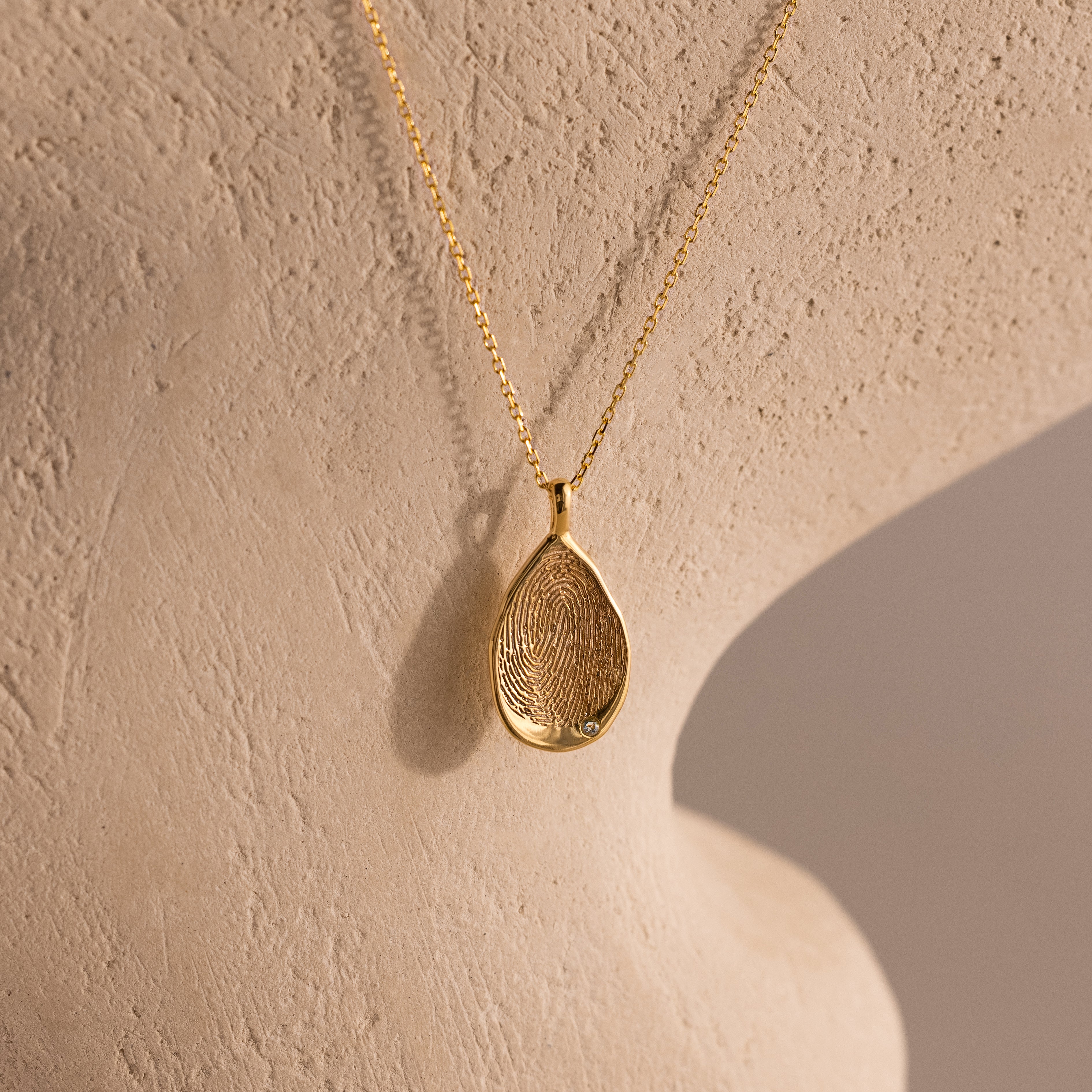 The Organic Fingerprint Teardrop Necklace in 18K Gold features a unique pendant, displayed on textured beige—a meaningful keepsake ideal for cherishing special memories.