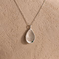 The Organic Fingerprint Teardrop Necklace in Sterling Silver features a textured teardrop and green gem on a delicate chain—a unique, personalized keepsake shown against a beige background.