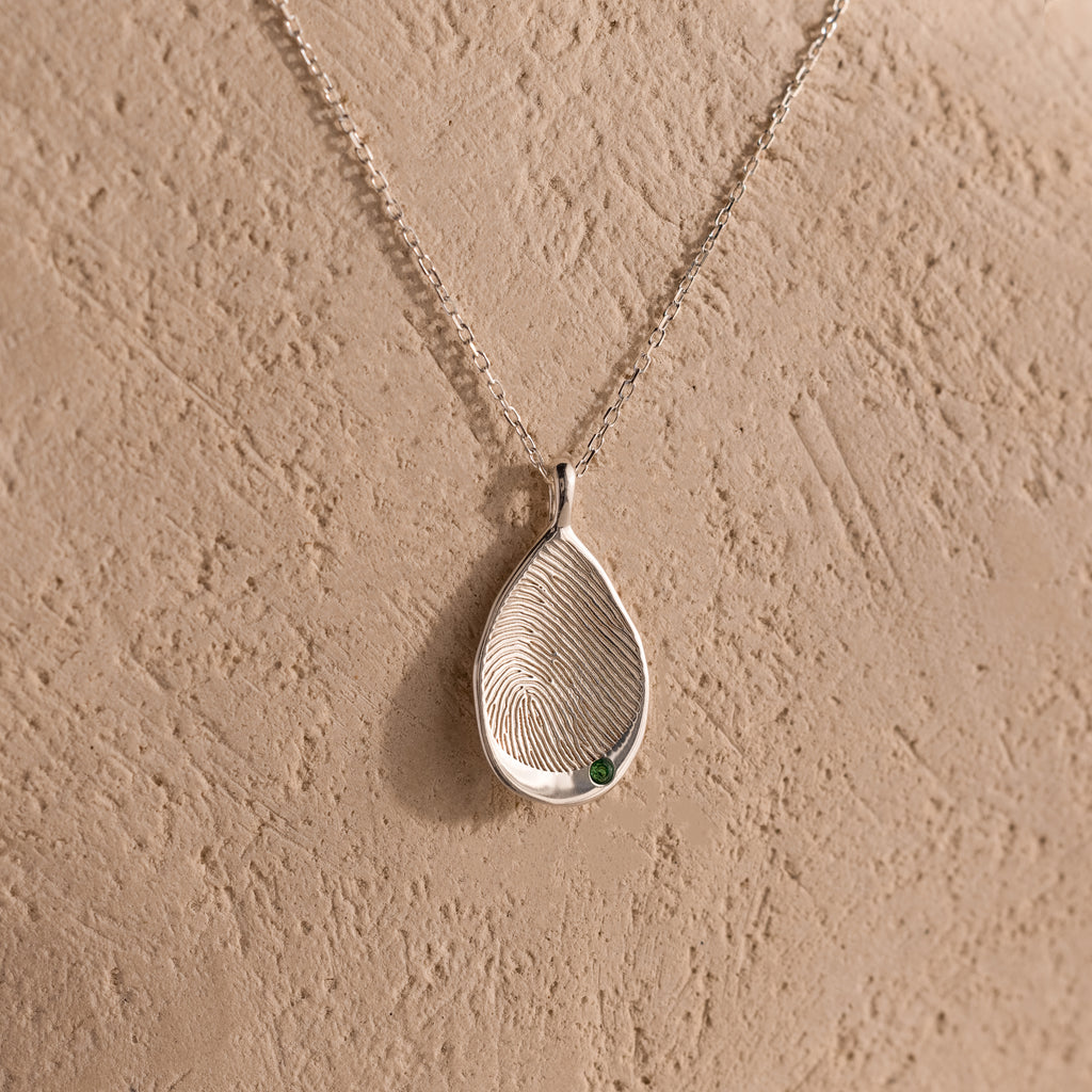 Organic Fingerprint Teardrop Necklace with a small green birthstone, displayed on a textured beige background—a personalized and uniquely charming jewelry piece.
