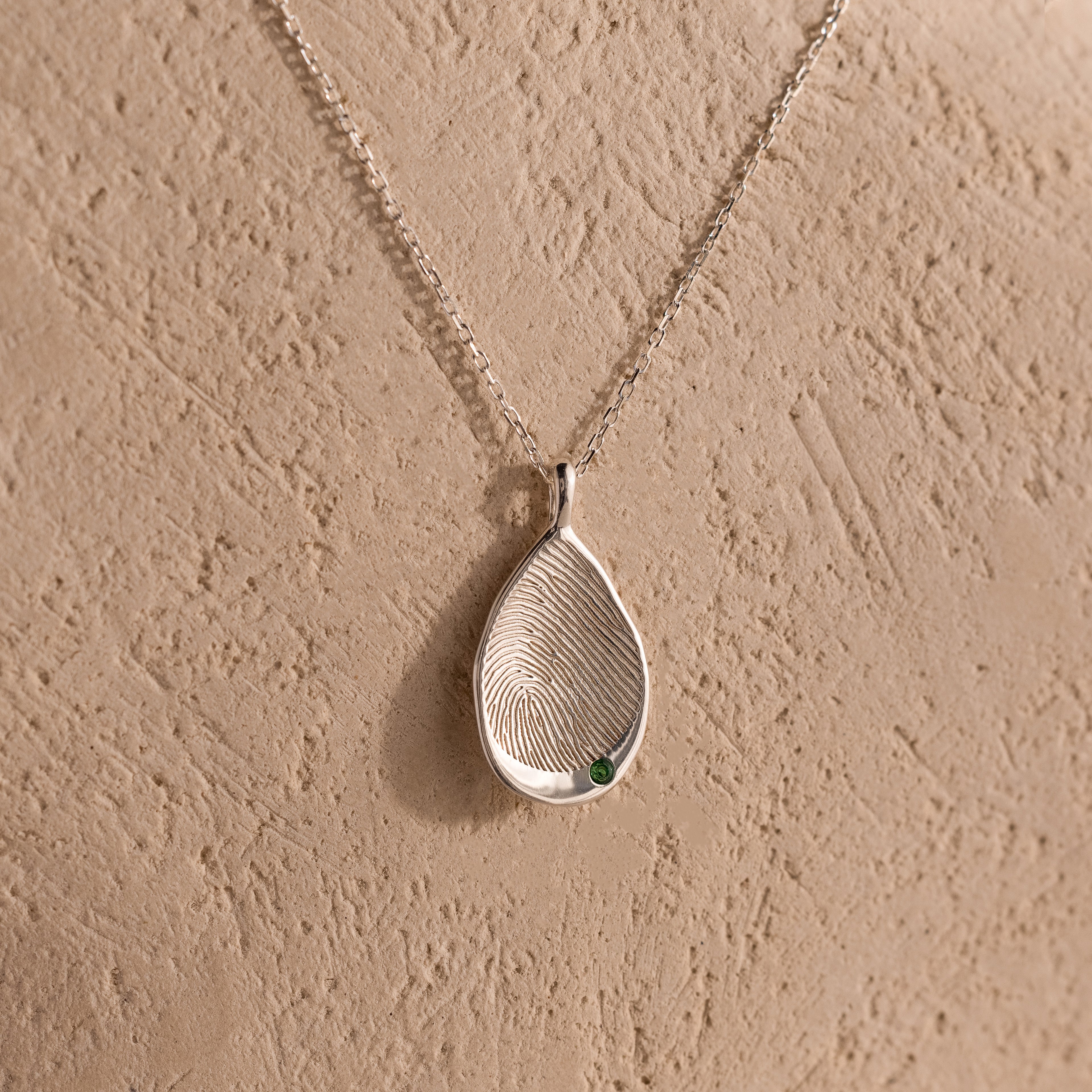 Organic Fingerprint Teardrop Necklace with a small green birthstone, displayed on a textured beige background—a personalized and uniquely charming jewelry piece.