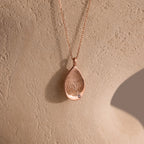 The Organic Fingerprint Teardrop Necklace in Rose Gold features a delicate diamond and is displayed on a textured surface for an elegant, organic look.