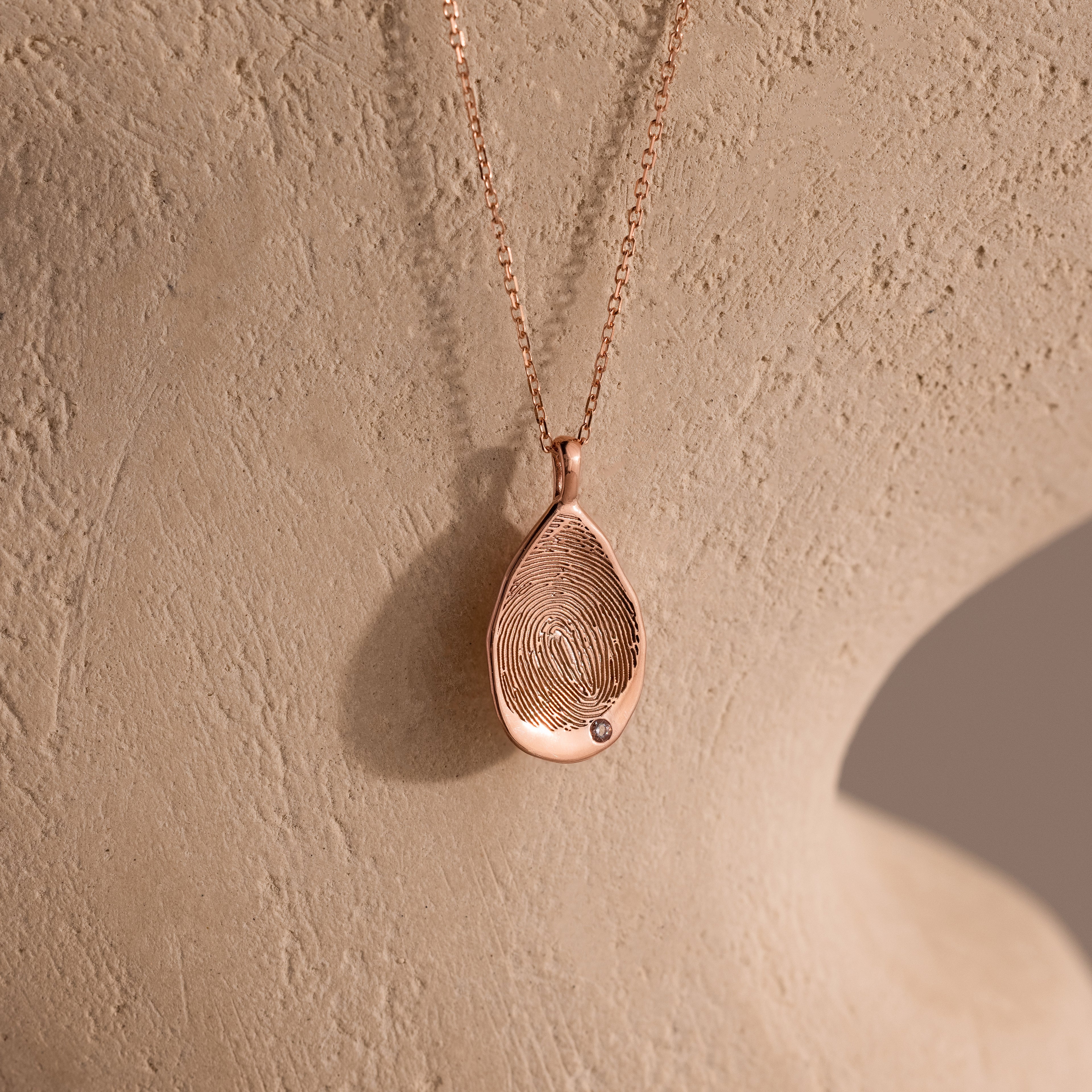 The Organic Fingerprint Teardrop Necklace in Rose Gold features a delicate diamond and is displayed on a textured surface for an elegant, organic look.