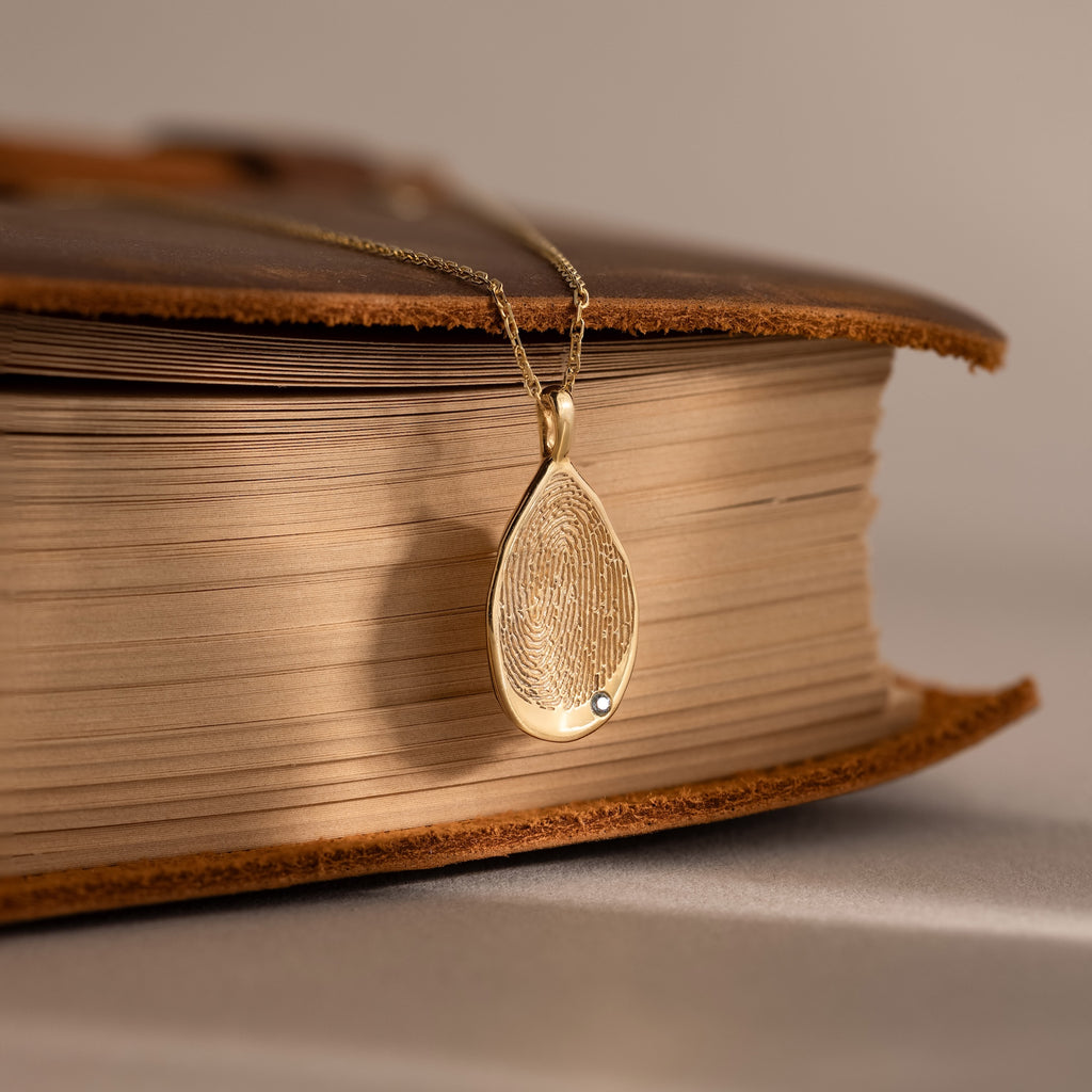 The Organic Fingerprint Teardrop Necklace in 18K Gold, featuring a delicate gem, is an elegant keepsake that sits gracefully atop a closed book.