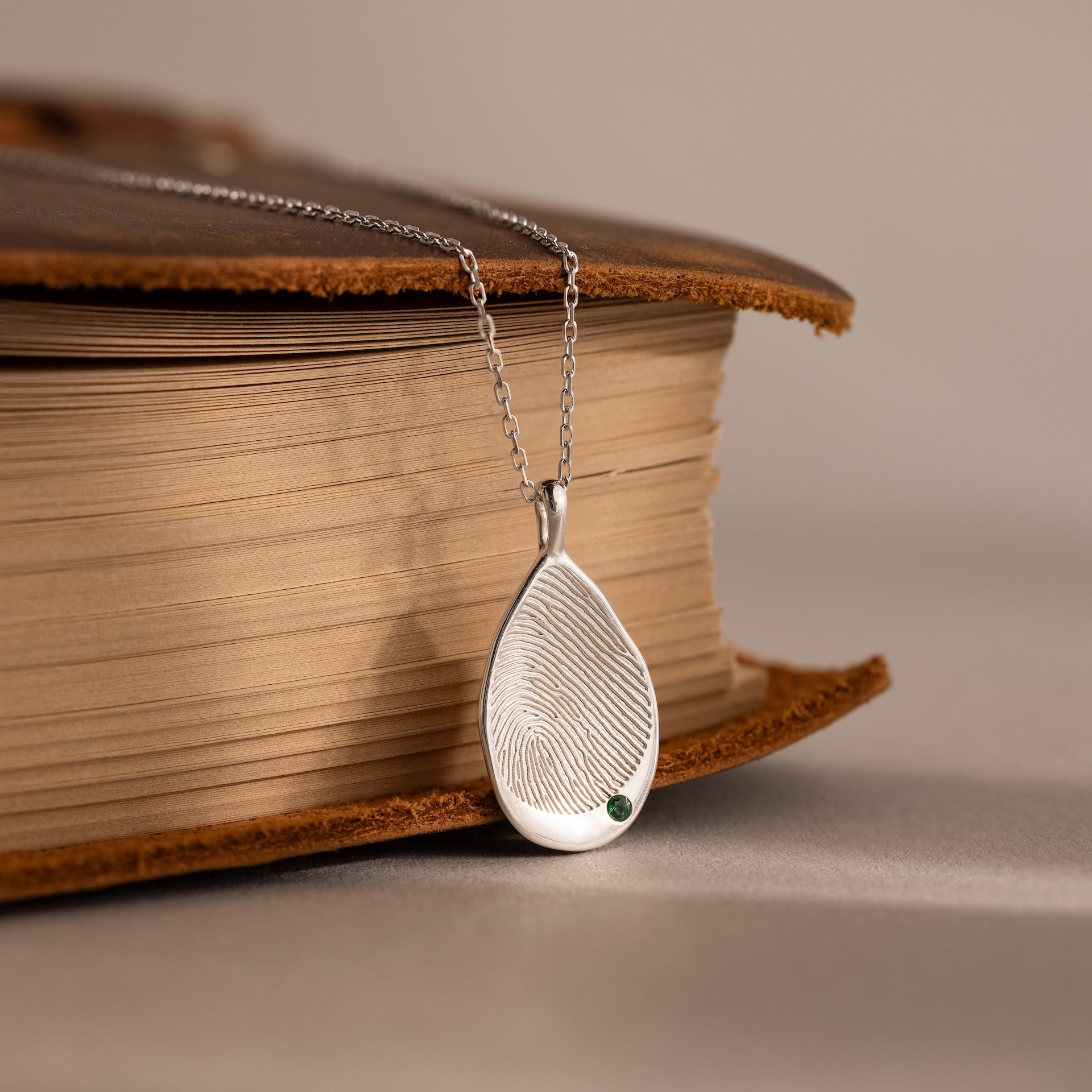 The Organic Fingerprint Teardrop Necklace in Sterling Silver, personalized as a birthstone necklace with a green gem, rests elegantly on a closed book—an unforgettable keepsake.