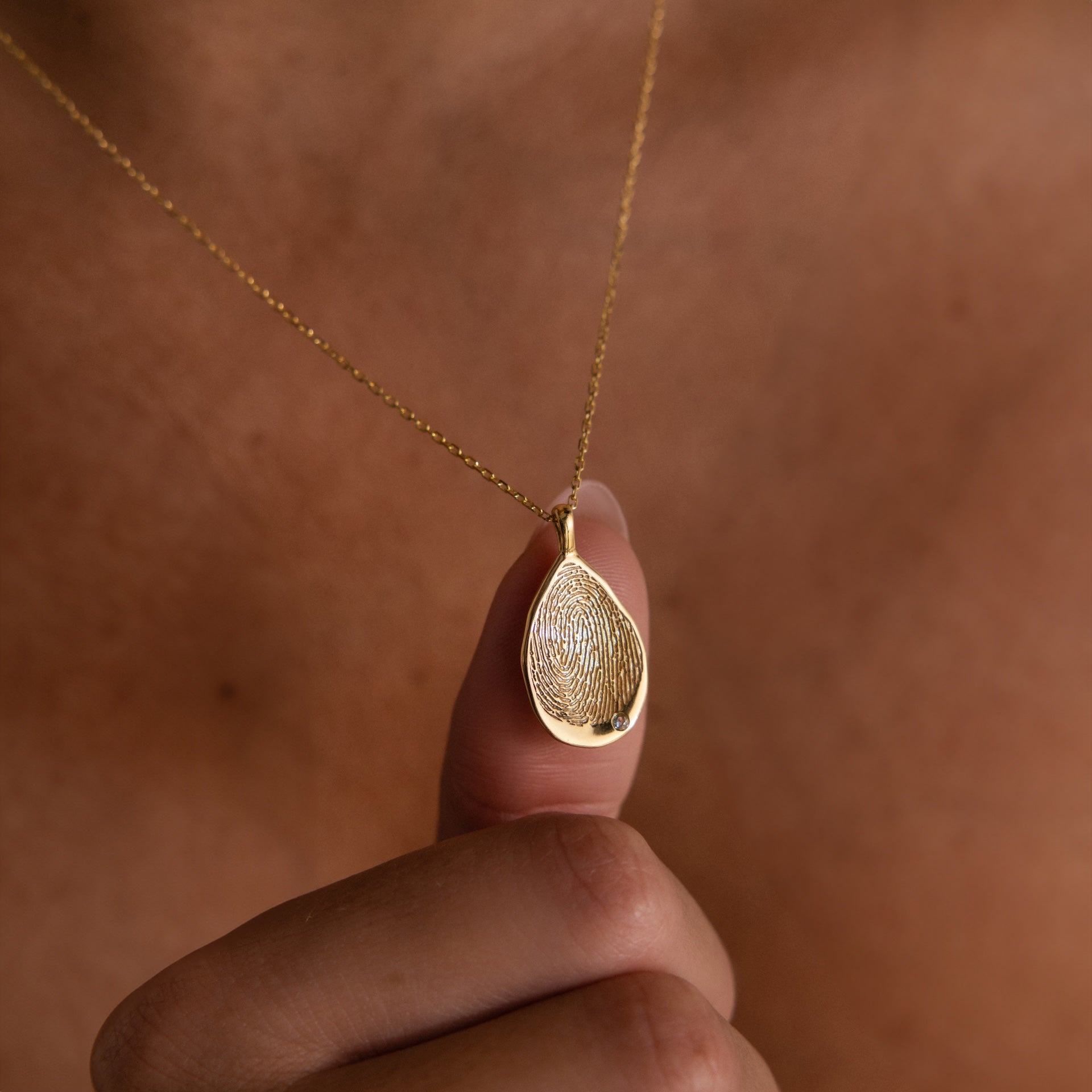 A person holds the Organic Fingerprint Teardrop Necklace in Rose Gold—a pendant engraved with a unique fingerprint.