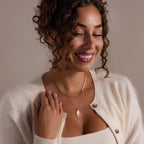 Smiling woman with curly hair, eyes closed, wears a cream sweater and the Organic Fingerprint Teardrop Necklace in 18K Gold.