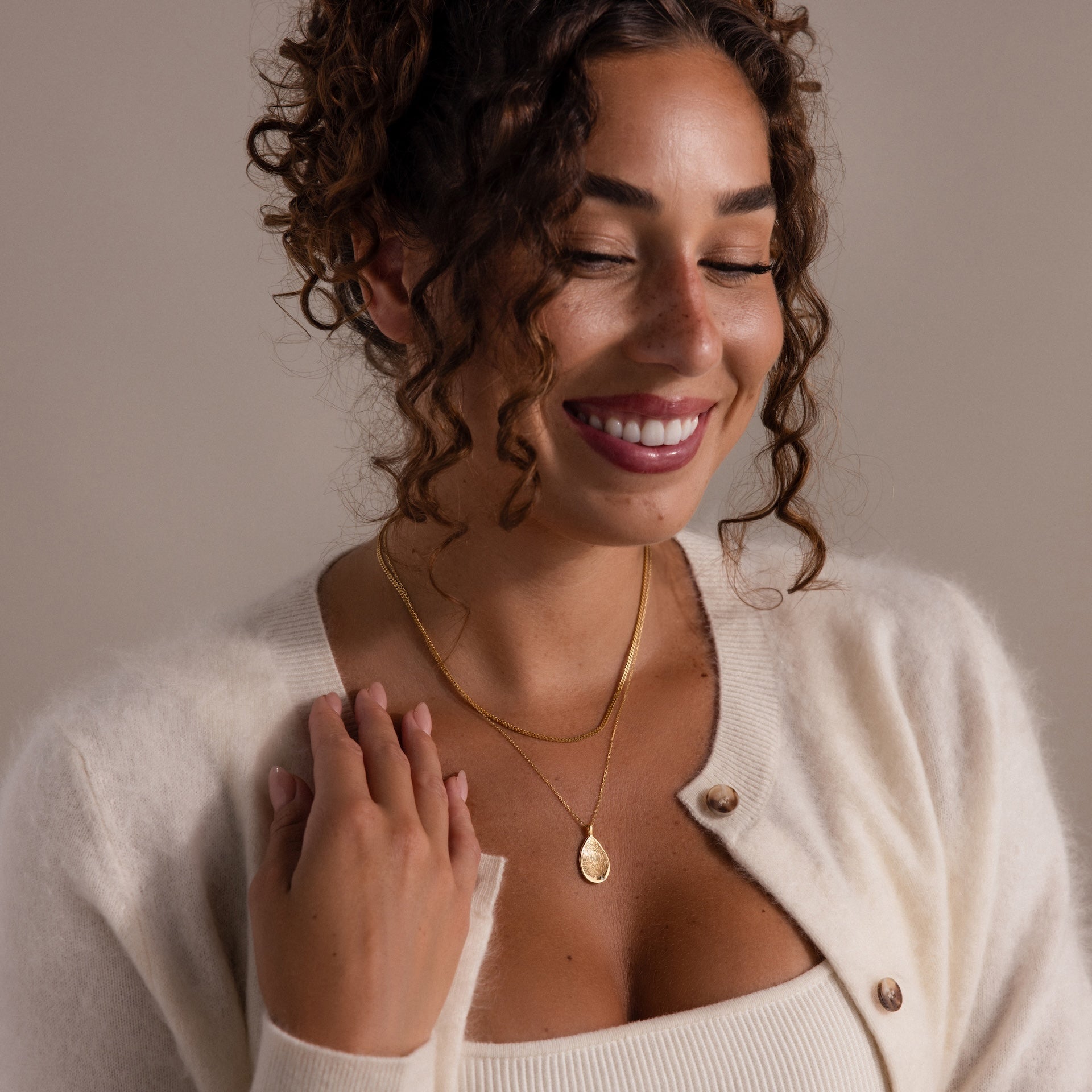 Smiling woman with curly hair, eyes closed, wears a cream sweater and the Organic Fingerprint Teardrop Necklace in 18K Gold.