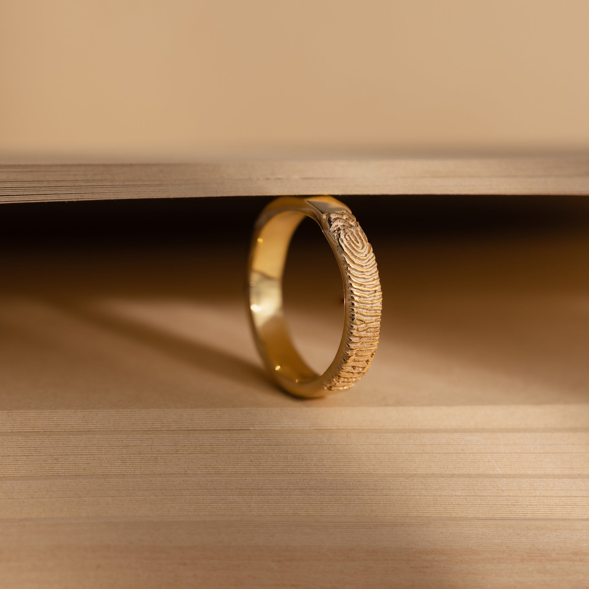 The Organic Fingerprint Band in 18K Gold stands upright between two wooden surfaces, its custom-engraved fingerprint pattern making it a unique memorial keepsake.