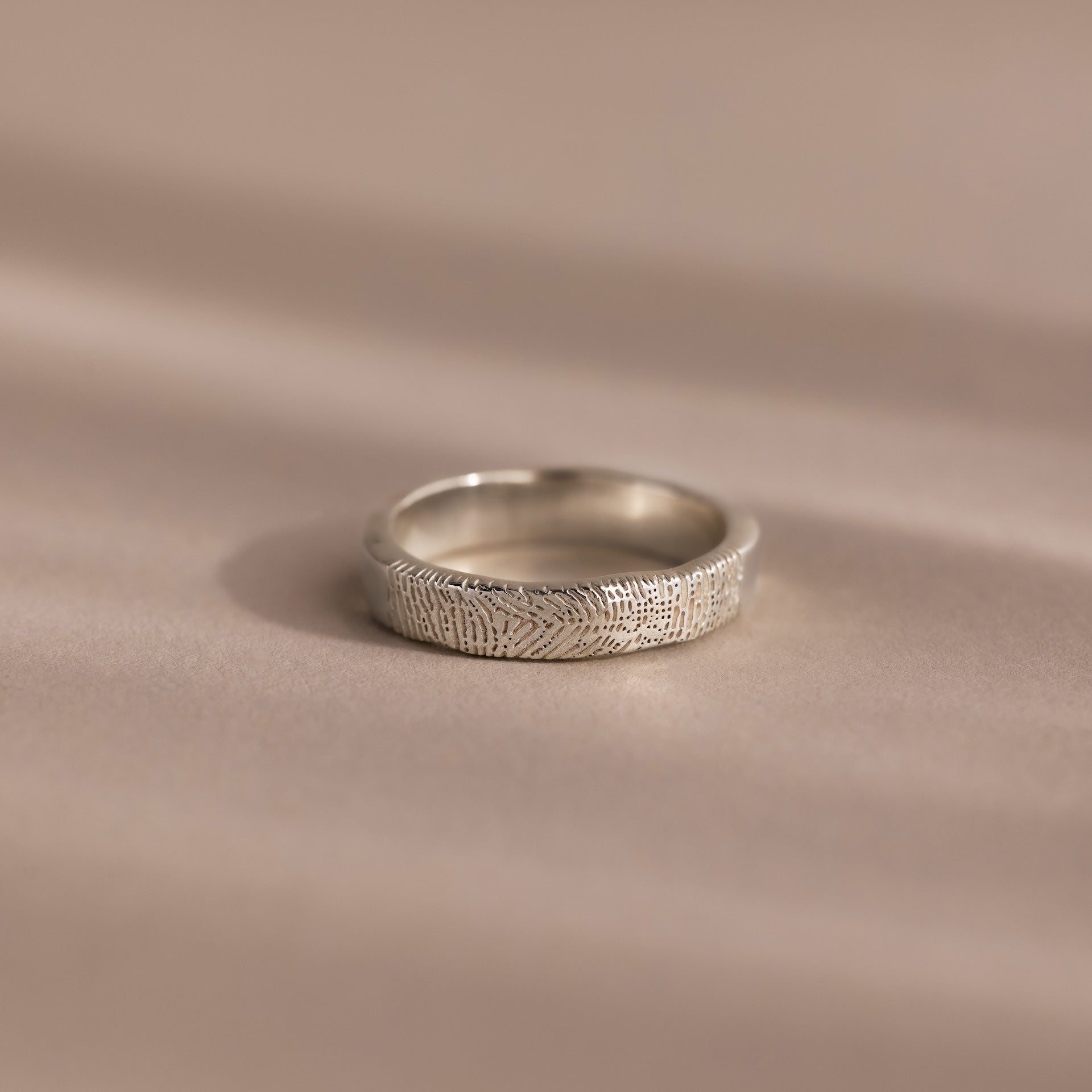 The Organic Fingerprint Band in Sterling Silver, featuring a delicate leaf pattern, rests on a soft beige surface—ideal as a memorial keepsake or custom-engraved ring for treasuring special memories.