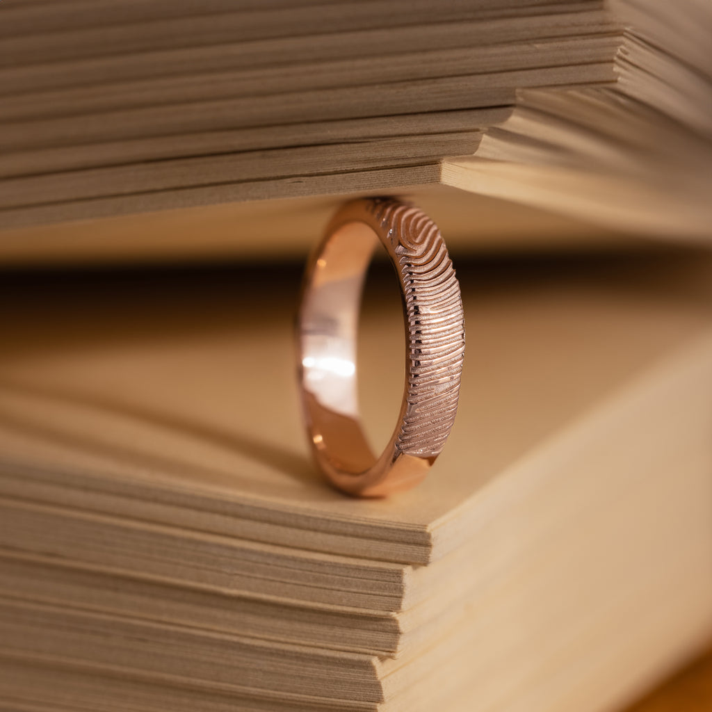 The Organic Fingerprint Band in Rose Gold stands upright between book pages, serving as a unique and memorable keepsake.