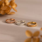 The Organic Fingerprint Band in Sterling Silver rests on a beige surface with blurred flowers in the background—a perfect custom-engraved memorial keepsake.