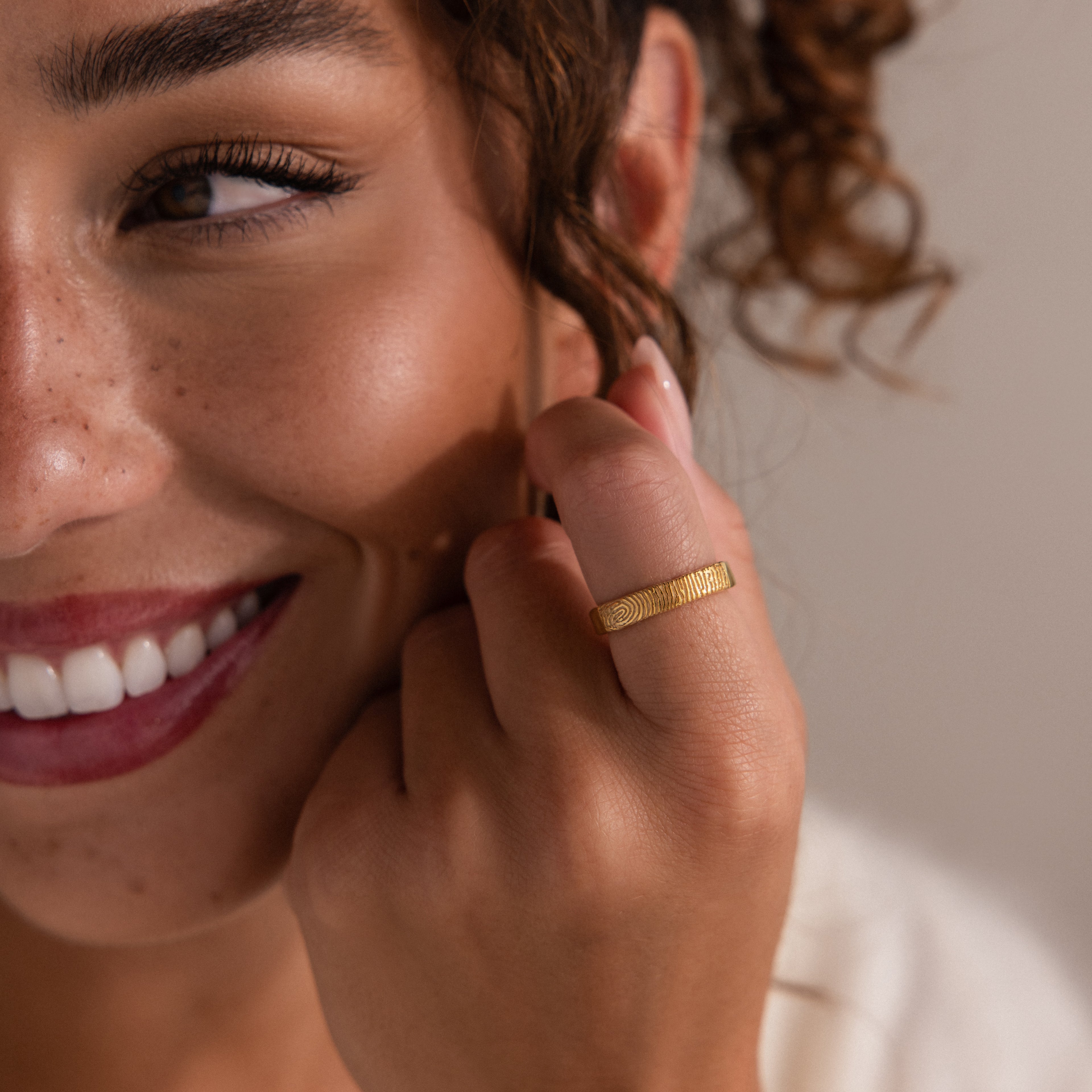 A woman with curly hair and freckled skin smiles, showing off the Organic Fingerprint Band in 18K Gold on her finger.
