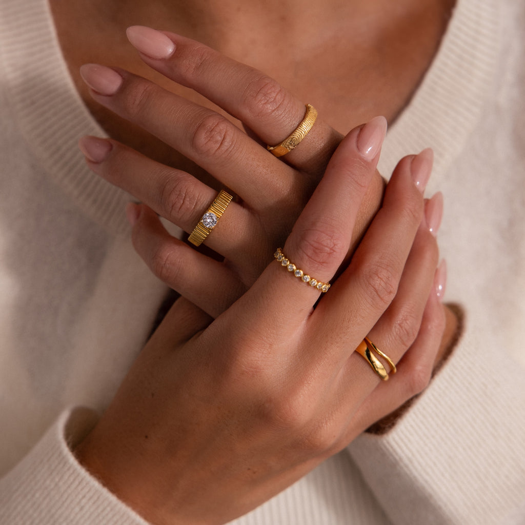 Close-up of hands with nude manicured nails and multiple gold rings, including the Organic Fingerprint Band, paired with a cream-colored sweater.